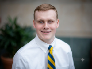 a smiling white man with short blonde hair wearing a white dress shirt and a yellow and blue striped tie against a blurred indoor background.