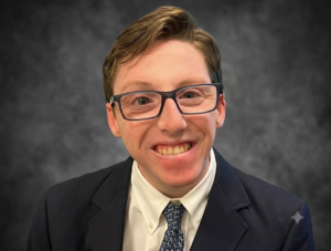 a white man with brown hair in a navy suit, wearing glasses, smiles at the camera.